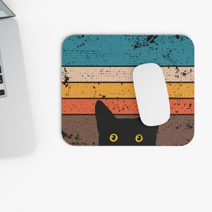 May include: A black cat with yellow eyes peeks out from behind a retro-style striped mouse pad. The mouse pad has a distressed, vintage look with faded blue, orange, yellow, and brown stripes.