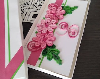 May include: A white gift box with a pink and green striped design. Inside, a handmade greeting card features pink paper flowers and green leaves. The card is decorated with paper quilling techniques.
