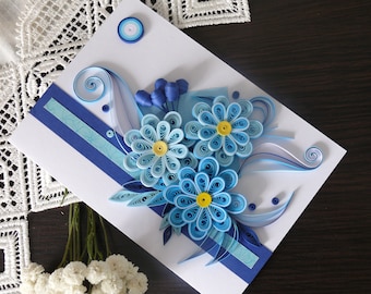 May include: A white card with a blue and white paper flower arrangement. The flowers are made of curled paper and have yellow centers. The card has a blue and white striped border.