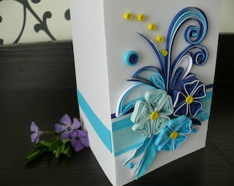 May include: A white card with a blue and turquoise paper flower arrangement. The flowers are made with quilled paper and have yellow centers. The card has a blue and turquoise ribbon around the bottom edge.