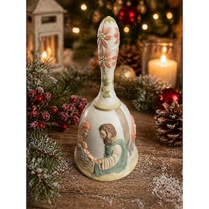 May include: A cream ceramic bell with a detailed handle featuring pink and green floral accents. The bell's body depicts a religious scene with figures in muted tones. The bell is set against a festive Christmas backdrop.