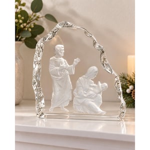May include: A clear glass sculpture featuring a nativity scene with three figures. The arched frame has a textured, organic edge. Inside, a standing figure and a seated figure holding a small infant are rendered in frosted white glass detail.