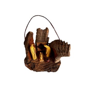May include: A rustic wooden nativity scene with carved figures of Mary, Joseph, and baby Jesus. The figures are light-colored against the dark brown wood base. A small, red, ceramic pot is also included in the scene. A dark wire loop is attached for hanging.