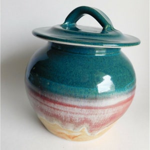 May include: A teal ceramic jar with a lid and a handle. The jar has a multi-colored glaze with shades of teal, pink, white, and yellow.