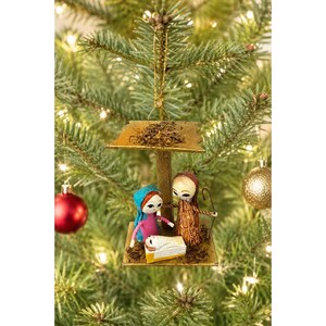 May include: A Christmas tree ornament depicting the Nativity scene. The ornament features a small wooden structure with figures of Mary, Joseph, and baby Jesus. The figures are dressed in colorful clothing. The ornament is hanging on a Christmas tree.