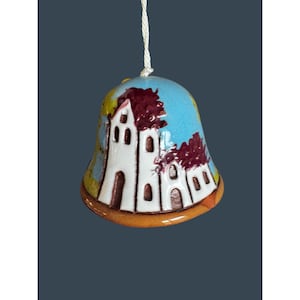 May include: A ceramic bell with a white church design, featuring brown roofs and arched windows, set against a blue sky background. The bell has a brown base and a white string for hanging.