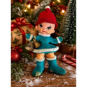 May include: A vintage-style figurine of a child wearing a bright red knit hat and a teal blue outfit with white faux fur trim. The figure has a light skin tone, painted facial features, and holds two small, textured gold-colored bells in one hand.