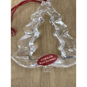 May include: Clear glass Christmas tree ornament with a red string for hanging. The ornament has a faceted design and a red oval sticker that reads "GORHAM West Germany".