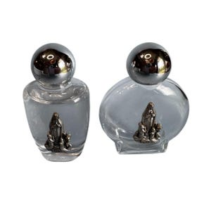 May include: Two clear glass bottles with silver-colored spherical caps. Each bottle has a silver-colored religious figure at the base. The bottles are filled with a light blue liquid. The word "FATIMA" is visible on the base of the figures.