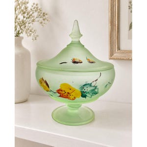 May include: A pale green frosted glass candy dish with a matching lid featuring a pointed finial. The bowl and lid are decorated with hand-painted autumn leaves in shades of yellow, orange, and teal, set on a circular pedestal base.