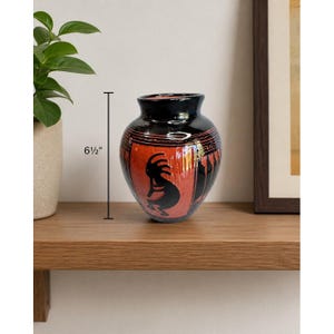 May include: A ceramic vase, approximately 6.5 inches tall, with a black rim and base. The vase features a red body with black Kokopelli figures and geometric designs. The vase is displayed on a wooden shelf.