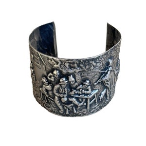 Pode incluir: Pulseira de prata com um design em relevo de um grupo de pessoas reunidas em torno de uma mesa. A cena se passa em um ambiente rústico ao ar livre com árvores e um edifício ao fundo.