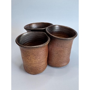 Puede incluir: Tres tazas de cerámica hechas a mano. Las tazas son de un color marrón rojizo cálido con un interior y borde marrón más oscuro. Las tazas tienen una forma ligeramente acampanada y un aspecto rústico y texturizado.