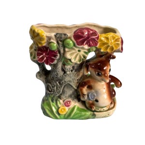 May include: A ceramic vase featuring a brown and white deer figure nestled beside a tree trunk. The tree is adorned with colorful flowers in shades of red, yellow, and green. The vase has a textured, rustic appearance, with a small opening at the top.