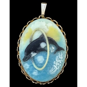 May include: Oval pendant with a gold-tone frame. It features a painted scene of a black and white orca jumping through a hoop. The background includes blue water, yellow and white clouds, and a gold-tone bail.