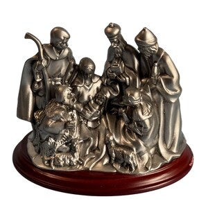 May include: A silver-colored nativity scene figurine depicting the three wise men, shepherds, and baby Jesus. The figures are arranged on a dark brown, round wooden base. The scene includes a lamb and other religious elements.