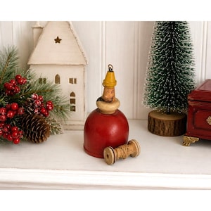 May include: A vintage wooden bell ornament featuring a bright red base, a cream-colored collar, and a small yellow pointed cap at the top. A small wooden spool wrapped with thin brown twine rests horizontally in front of the red bell base.