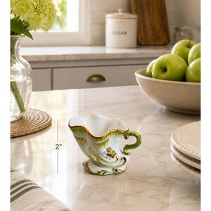 May include: A small white ceramic creamer with a green scalloped rim, gold accents, and a painted butterfly motif. The creamer features a curved handle and a flared spout, with a visible measurement indicator showing a height of 2 inches.