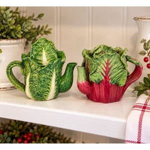 May include: Two ceramic teapots shaped like vegetables. The left teapot resembles a head of green lettuce with textured leaves. The right teapot resembles a bunch of Swiss chard with green leaves and prominent red stalks and veins.