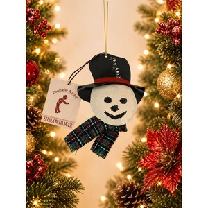 Peut inclure: Une décoration de bonhomme de neige avec un haut-de-forme noir à bande rouge, un visage blanc aux traits peints en noir et une écharpe en tissu à carreaux verts, bleus et rouges. Une étiquette beige porte le texte Decorative Accents Shadowdancer.