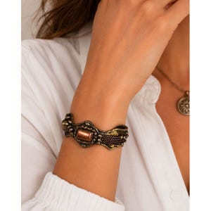 Op de afbeelding: Een bronzen manchetarmband met een rechthoekige koperkleurige middensteen. De armband heeft ingewikkelde details, waaronder een geweven gedeelte en decoratieve versieringen. Een bijpassende hangerketting is ook zichtbaar.