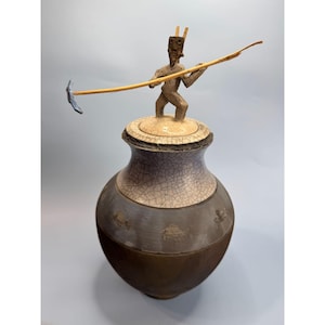 Puede incluir: Un jarrón de cerámica con un esmalte craquelado y un diseño marrón y blanco. El jarrón tiene una tapa con una pequeña figura de una persona sosteniendo un palo.