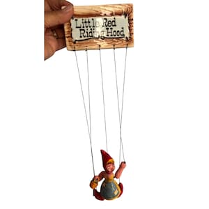May include: A vintage Little Red Riding Hood marionette doll. The doll is suspended from a wooden sign with the text "Little Red Riding Hood". The doll is wearing a red hat, yellow top, blue skirt, and red shoes. The doll is holding a basket.