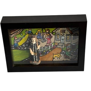 May include: A framed folk art piece featuring a painted village scene with figures and a central figurine. The artwork is set within a black rectangular frame. The scene includes a house, a dog, and people engaged in various activities.