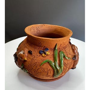 Può includere: Un vaso in ceramica color terracotta con un'ampia apertura e una base rotonda. Il vaso è decorato con rilievi di piante, una farfalla e piccoli animali. Il testo "June Sears" è visibile sul bordo.