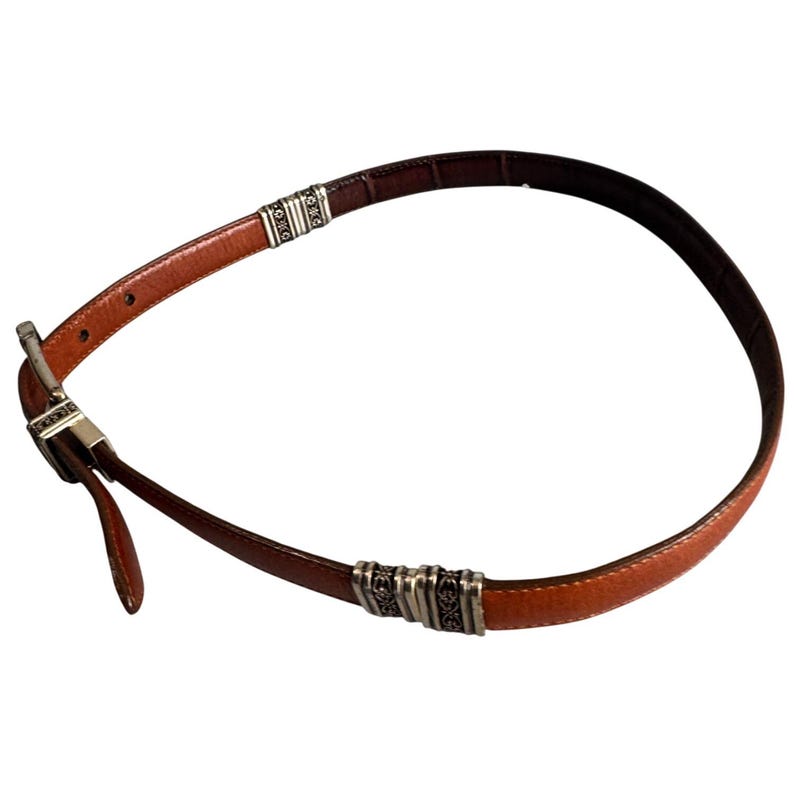 Peut inclure: Une ceinture en cuir marron dot&eacute;e d'une boucle en m&eacute;tal argent&eacute; et de deux ornements d&eacute;coratifs en m&eacute;tal argent&eacute; le long de la sangle. La ceinture pr&eacute;sente une finition lisse &agrave; l'ext&eacute;rieur et une doublure int&eacute;rieure marron fonc&eacute; avec des coutures.