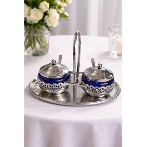 May include: A silver-toned condiment set with two blue glass jars, each with a silver lid and spoon. The jars sit on a silver tray with a handle. The jars have a decorative silver filigree around the base.