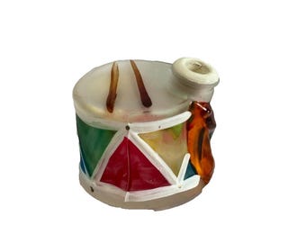 May include: A colorful, miniature drum ornament with a white base and a cylindrical shape. The drum features a red, green, and yellow design, with brown drumsticks on top. A white spout and an orange accent complete the festive look.