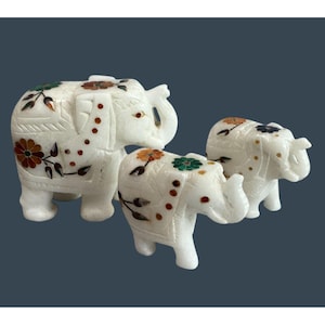May include: Three white marble elephant figurines with floral and dot inlay in shades of red, orange, green, and black. The elephants are in different sizes, with the largest in the front.