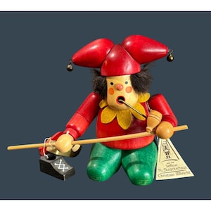 May include: A wooden jester figurine with a red hat, red top, and green pants. The jester has a yellow collar, black hair, and a wooden pipe. The figurine is holding a wooden stick and a small wooden drum. A tag reads "Seiffener Nußknackerhaus Christian Ulbricht."