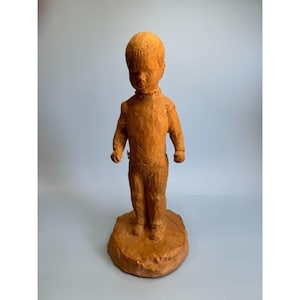 Könnte beinhalten: Eine Terrakotta-Skulptur eines Kindes, das auf einem Sockel steht. Die Skulptur ist aus Papiermaché gefertigt und hat einen rustikalen, handgefertigten Look.