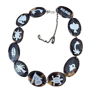 Op de afbeelding: Een ketting met ovale kralen in een schildpadpatroon. Elke kraal heeft een wit ontwerp, waaronder een zeilboot, een schildpad en een palmboom. De ketting heeft een zilverkleurige ketting en sluiting.
