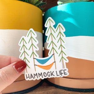 Może przedstawiać: Biała naklejka z hamakiem między dwoma drzewami. Naklejka zawiera napis "HAMMOCK LIFE" czarnym tekstem.