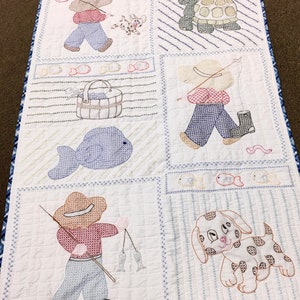 May include: A white quilted baby blanket with embroidered images of a boy fishing, a dog, a turtle, and a fish. The blanket has a blue and white checked border.