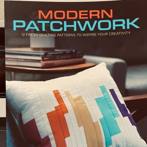 May include: A white pillow with a colorful patchwork design featuring rectangular blocks in shades of purple, orange, gold, teal, and blue. The book cover in the background reads "MODERN PATCHWORK" and "12 FRESH QUILTING PATTERNS TO INSPIRE YOUR CREATIVITY."