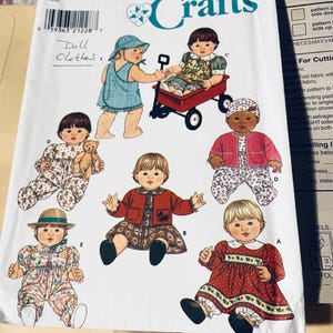 May include: A Simplicity Crafts sewing pattern envelope, number 7992, featuring illustrations of dolls wearing various outfits including dresses, hats, and jackets. The white paper envelope displays the text Doll Clothes handwritten in blue ink.