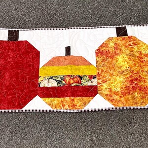 May include: A quilted table runner with five stylised pumpkins in orange, yellow, and red. The pumpkins are set against a white background with a black and white checkered border. The runner is approximately 122 cm long.