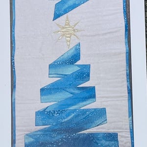 May include: A quilted Christmas tree design with a light gray background. The tree is made of blue ribbon-like shapes, with a gold star at the top. The edges of the quilt are bordered with a darker gray fabric.