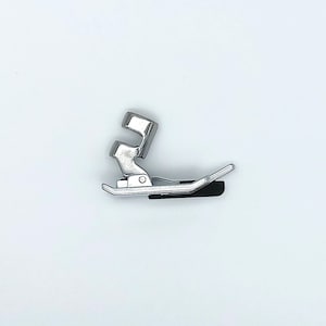 May include: A silver-colored sewing machine presser foot, designed for various sewing tasks. The foot has a curved shape with a black attachment piece. The image is set against a plain white background, highlighting the metallic sheen of the foot.
