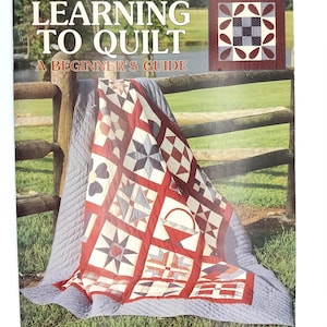 以下が含まれることがあります： Lori Yetmar Smith著のソフトカバーの教本「Learning to Quilt: A Beginner's Guide」。表紙には、様々な幾何学模様が施された赤、白、青のパッチワークキルトと、25のレッスンを示す小さな画像が掲載されています。