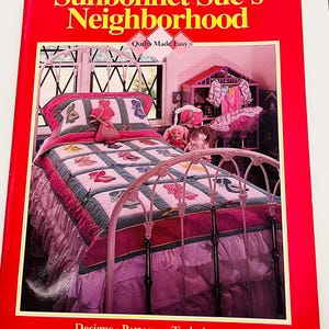 May include: A softcover book titled Sunbonnet Sue's Neighborhood by Quilts Made Easy. The cover features a photograph of a twin-sized bed with a pink metal frame, a patchwork quilt with Sunbonnet Sue motifs, and a pink ruffled bed skirt.