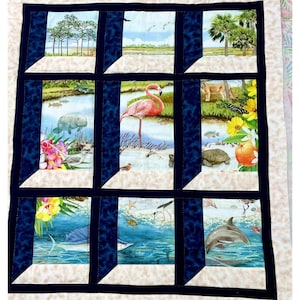 May include: A quilt with a windowpane design featuring various Florida wildlife, including a manatee, a flamingo, a panther, a turtle, a marlin, and a dolphin. The quilt is made with a beige background and blue window frames.