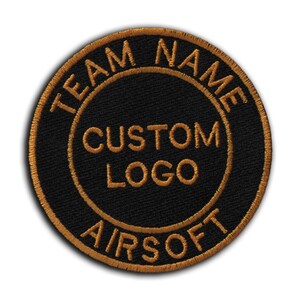 Puede incluir: Parche redondo negro con texto bordado en dorado. El texto dice "TEAM NAME" en la parte superior, "CUSTOM LOGO" en el centro y "AIRSOFT" en la parte inferior. El parche tiene un borde dorado.