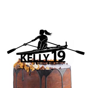 Peut inclure: Décoration de gâteau noire représentant une silhouette d'une personne ramant dans un bateau, avec le nom "KELLY" et le chiffre "19" découpés en dessous. Le gâteau est décoré de glaçage au chocolat et de garnitures.