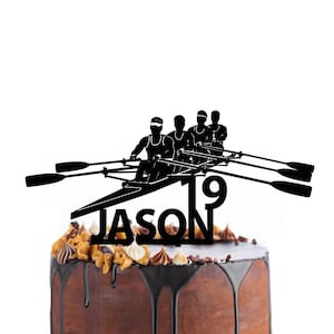 以下が含まれることがあります： ボート競技チームのシルエット、名前「JASON」、数字「19」が描かれた黒いケーキトッパー。トッパーは、装飾的なトッピングとチョコレートのドリップが付いたチョコレートケーキの上に置かれています。