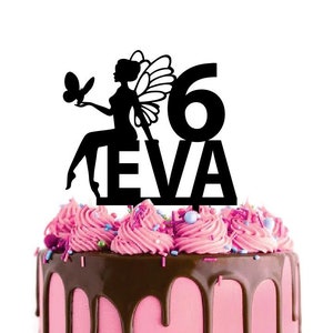 Peut inclure: Décor de gâteau en silhouette noire représentant une fée avec des ailes, un papillon et le chiffre 6. Le nom "EVA" est écrit sous le chiffre. Le décor est sur un gâteau rose et brun avec un glaçage au chocolat.